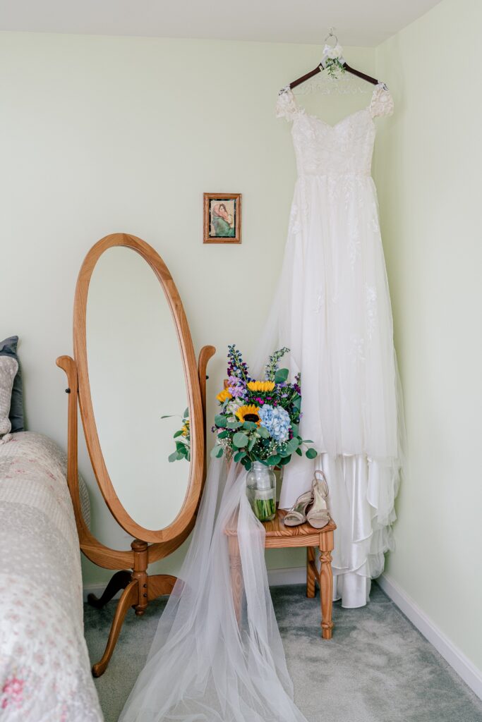 A wedding dress hanging beside a mirror with a bouquet and shoes on a chair beside it