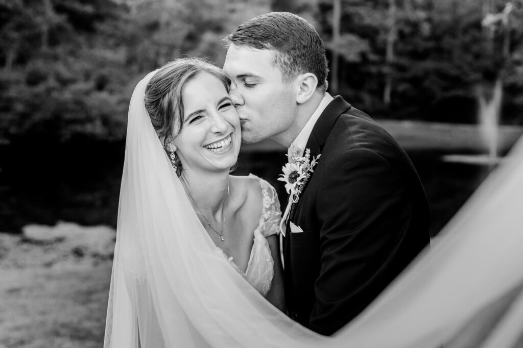 A bride laughs at the camera as her groom kisses her cheek