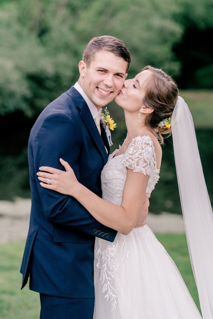 A bride kisses the cheek of her groom as he smiles at the camera