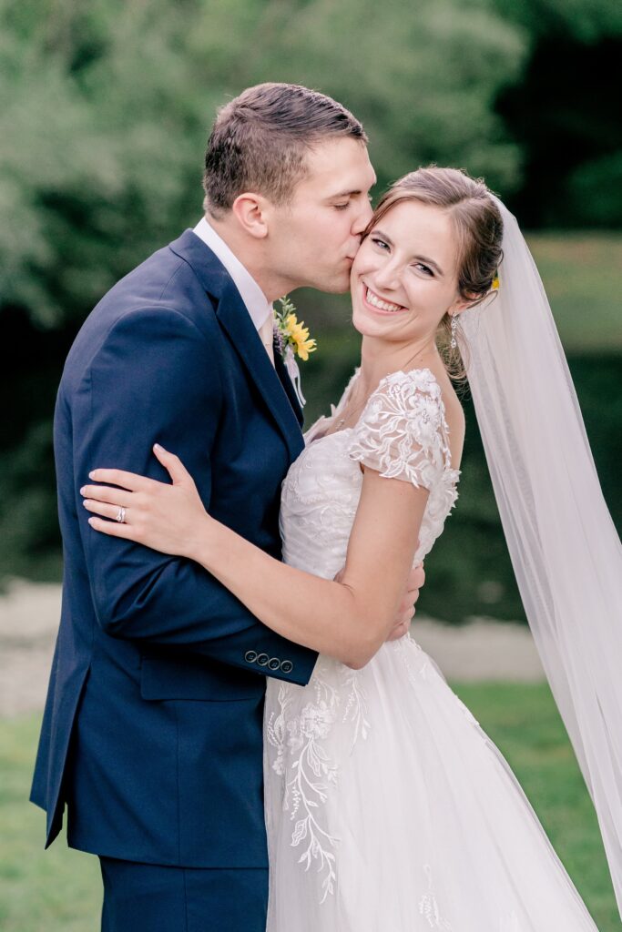 A bride smiles at the camera as her groom kisses her cheek during their Stevenson Ridge wedding reception