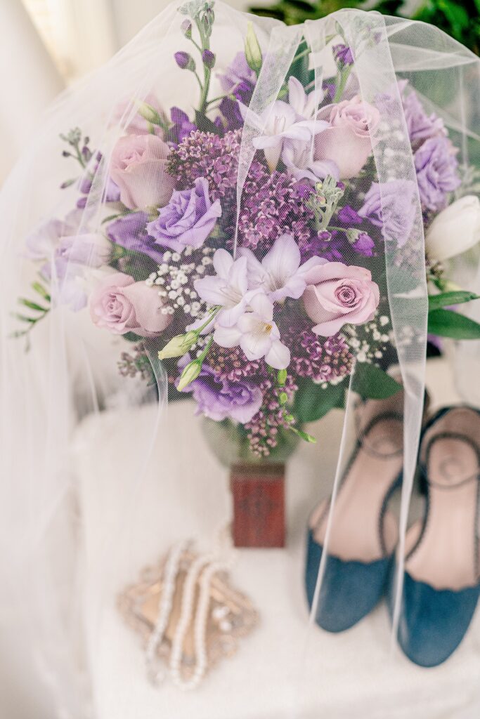 A bridal bouquet of purple flowers beside the bride's jewelry and shoes before her Catholic wedding