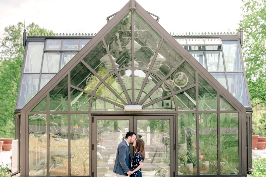 A couple shares a kiss in front of the greenhouse during their Meadowlark Botanical Gardens engagement session