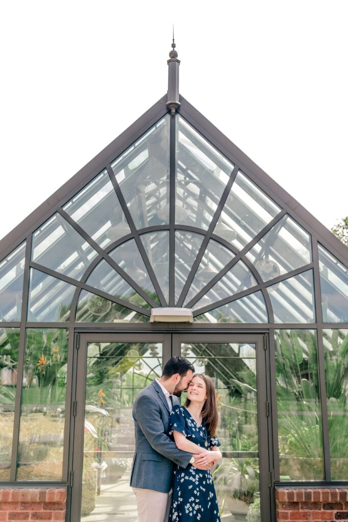 A couple snuggles in front of the greenhouse during their Meadowlark Botanical Gardens engagement session