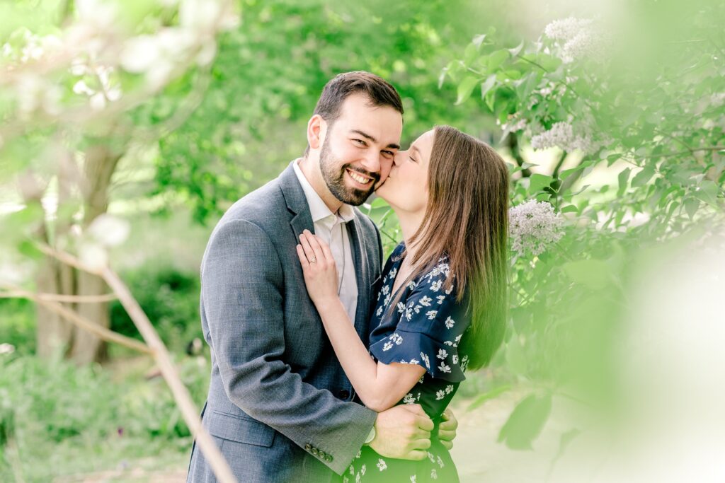 A woman kisses her groom-to-be on the cheek among flowers during their Meadowlark Botanical Gardens engagement session