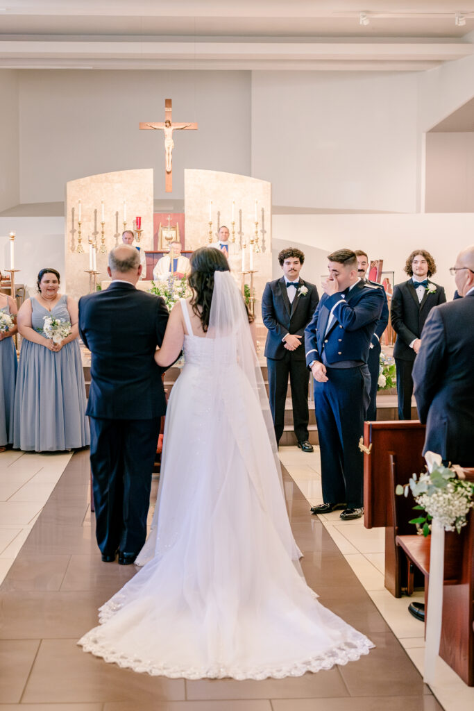 A groom wiping tears as his bride processes down the aisle during a wedding at St Philip Catholic Church in Falls Church Virginia