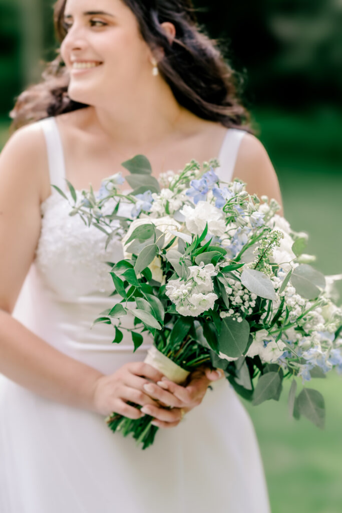 A bride smiles over her shoulder while holding a bouquet of white and blue flowers during a tented wedding reception at Historic Blenheim in Fairfax, Virginia