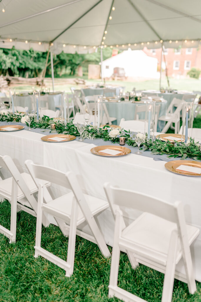 The head table featuring soft blue taper candles and gold chargers surrounded by greenery and white roses during a tented wedding reception at Historic Blenheim in Fairfax, Virginia