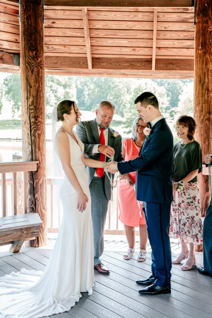 A bride and groom laughing as their family member ties a knot around their hands