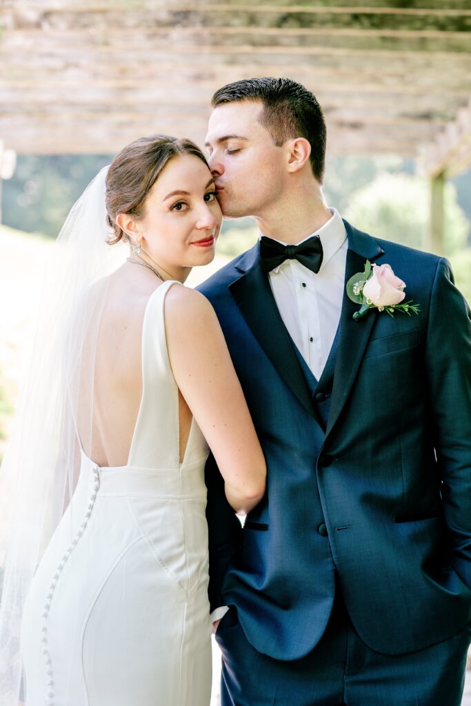 A groom kisses his bride on the temple as she looks over her shoulder at the camera