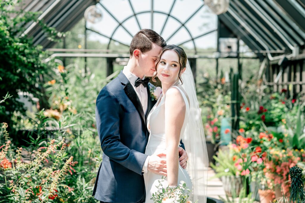 A bride looks over her shoulder as her groom nuzzles into her inside a greenhouse 