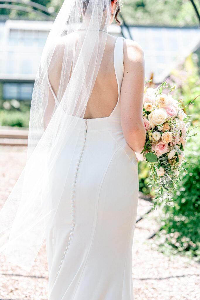 Elegant buttons down the back of a bride's wedding dress as she holds her bouquet and her veil flows off to the side