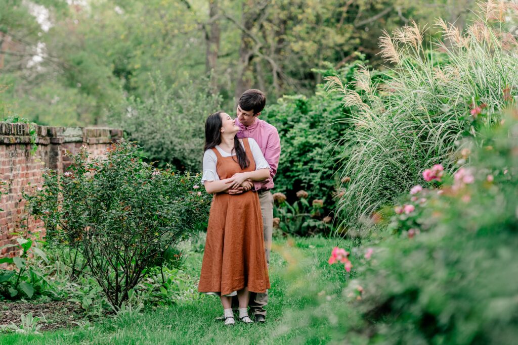 An engaged couple look at each other and smile as he wraps his arms around her waist in a rose garden