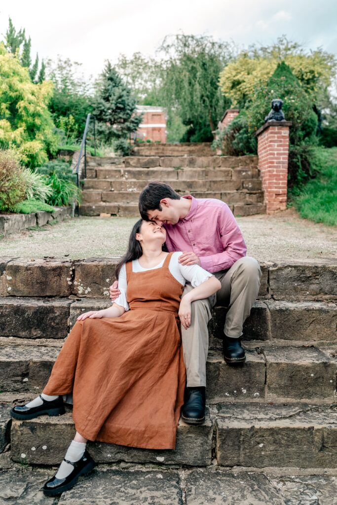 An engaged couple smiling forehead to forehead as they sit on a grand staircase in a historic garden