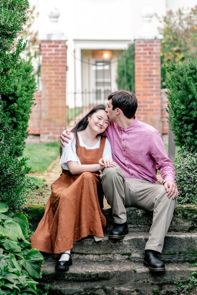 A man kisses the forehead of his fiancée while sitting on a stone staircase in a curated garden