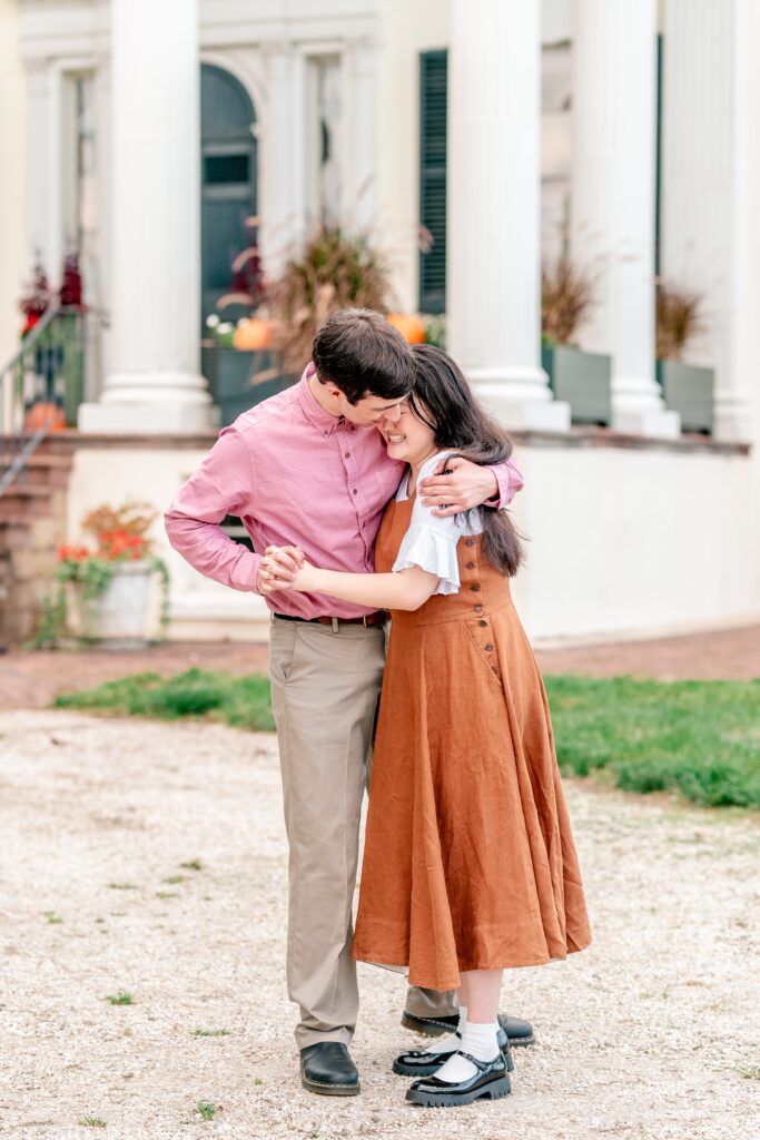 An engaged couple laughs together as they dance in front of a historic house