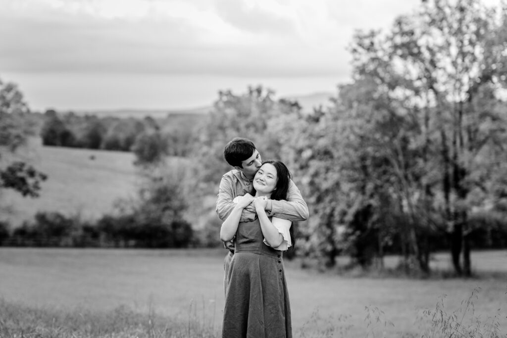 A man kisses the side of his fiancée’s head with his arms wrapped around her from behind and she smiles at the camera while they stand in an open field