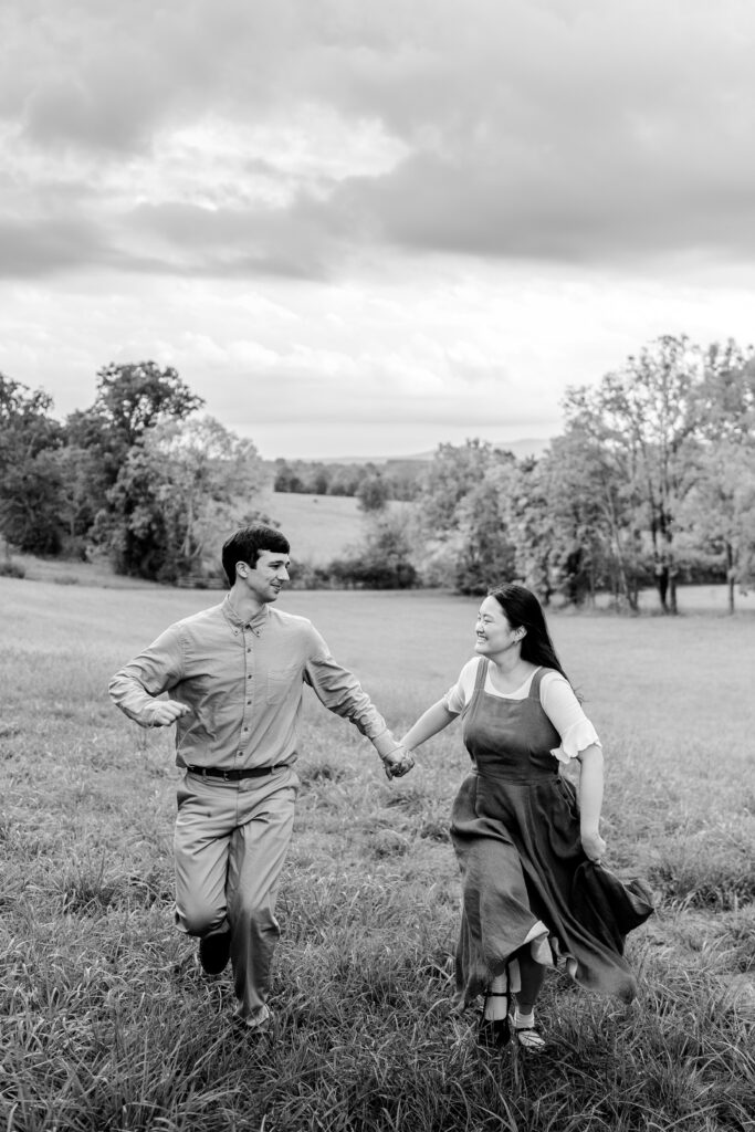 An engaged couple holds hands and runs through an open field together