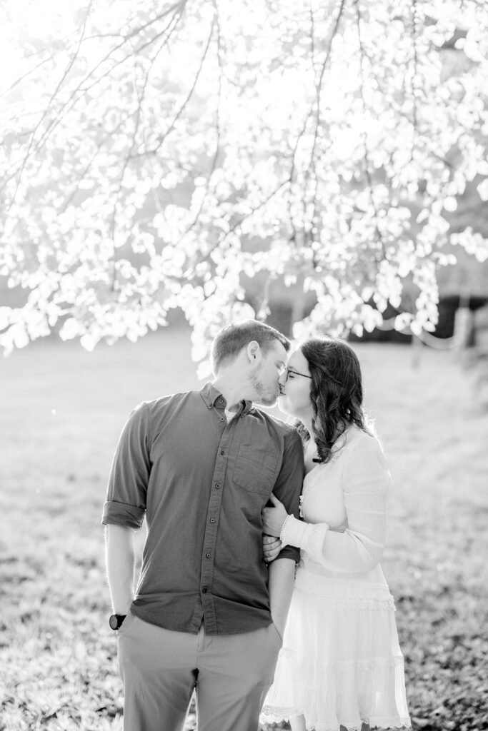 A black and white image of an engaged couple sharing a kiss under a tree