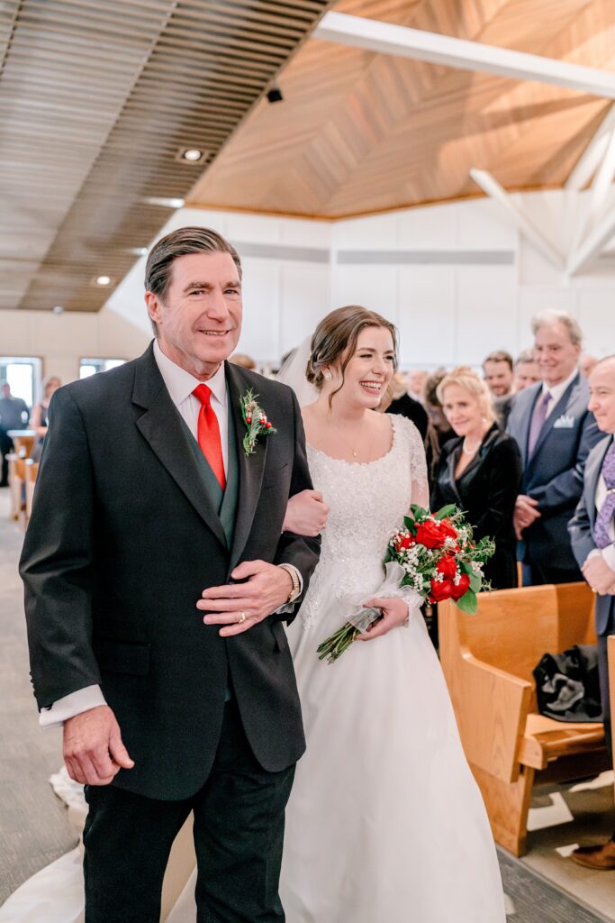 A father smiles at the camera as he walks his daughter down the aisle during a Christmas themed wedding at St. Joseph Catholic Church in Herndon Virginia