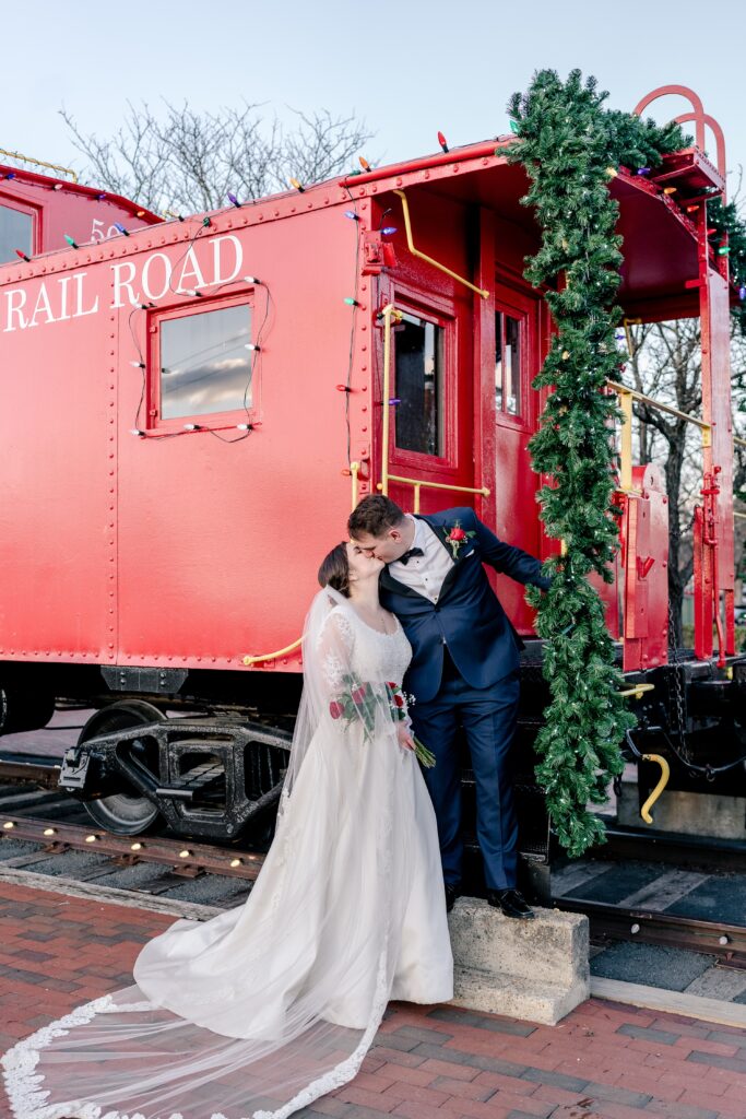 A bride and groom share a kiss next to a red train car decorated for Christmas during their wedding in Herndon VA