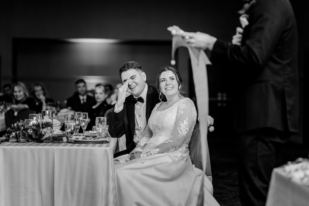 A groom laughs as his friend gives a toast at his wedding