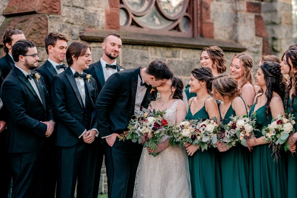 A bride and groom share a kiss surrounded by their wedding party