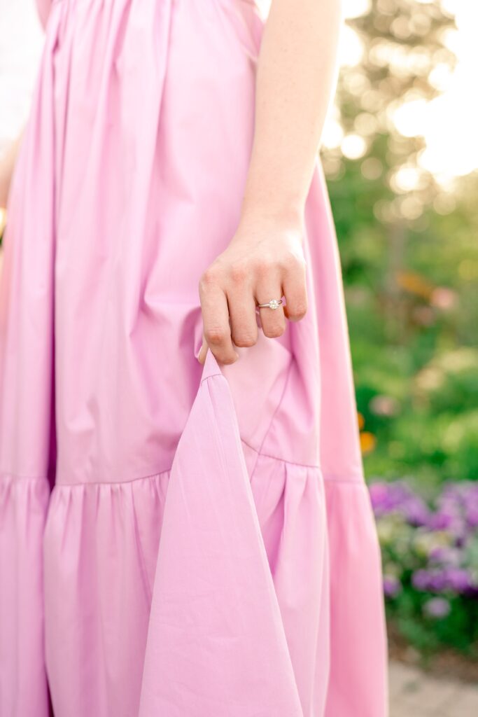 A close up of a woman's hand with a circle cut engagement ring holding her pink skirt