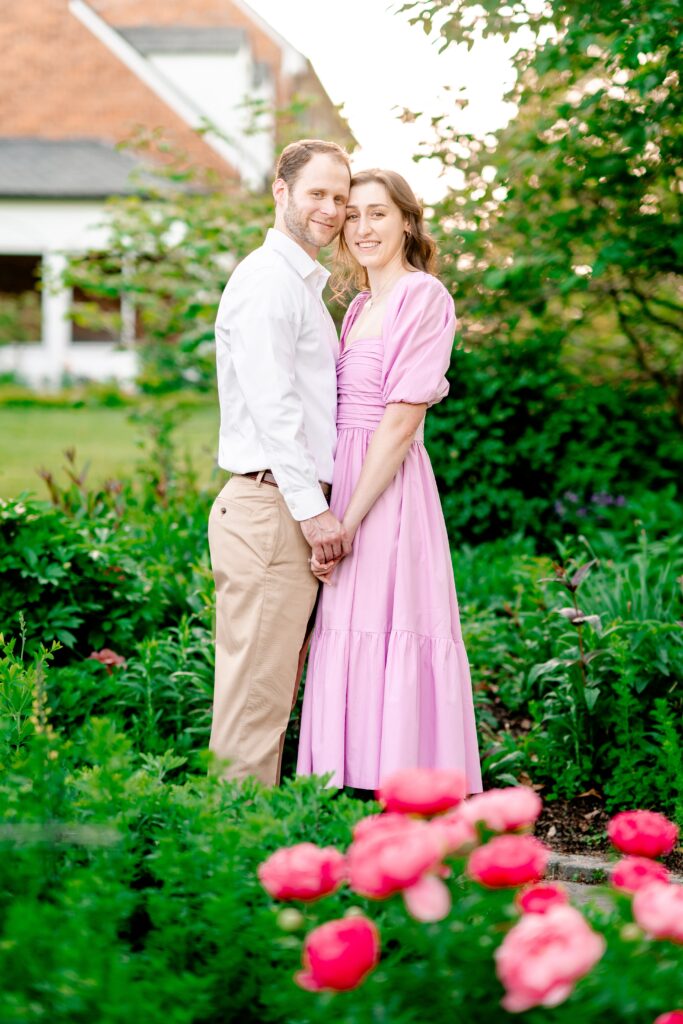 An engaged couple posing for a classic portrait in front of a historic house and peony bush