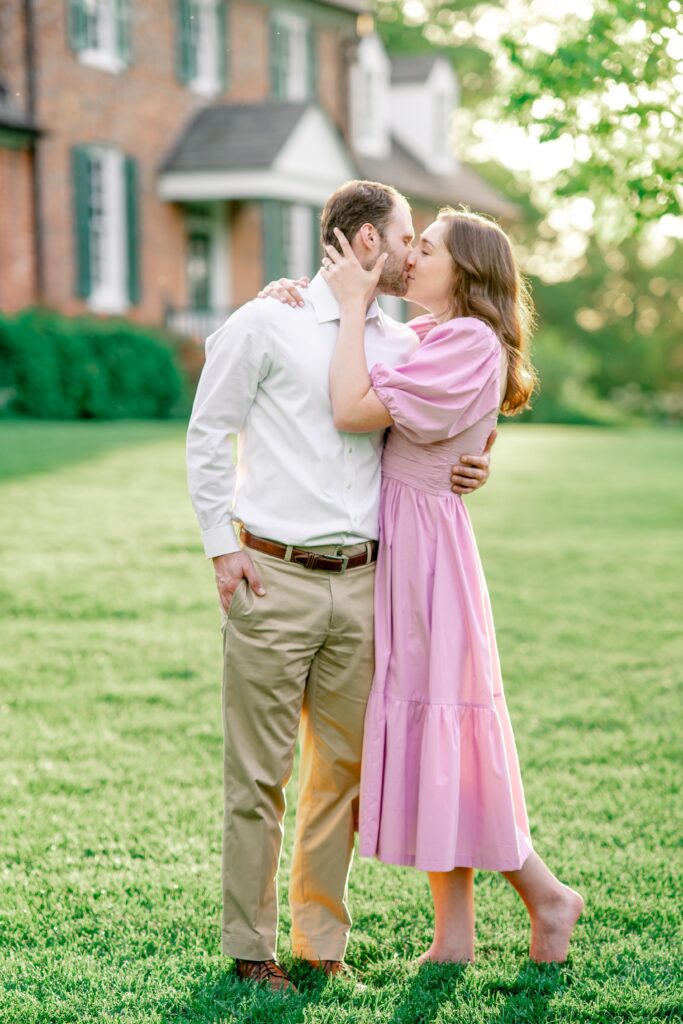 An engaged couple shares a kiss in front of a historic house