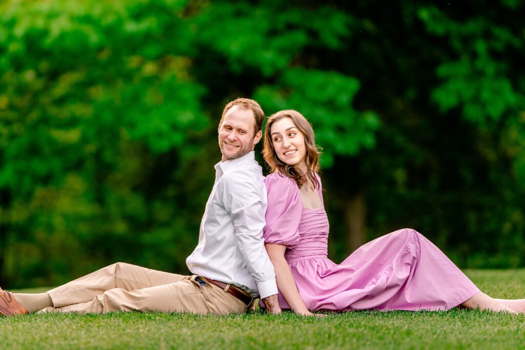 An engaged couple sits on the grass together back to back and look at each other over their shoulders