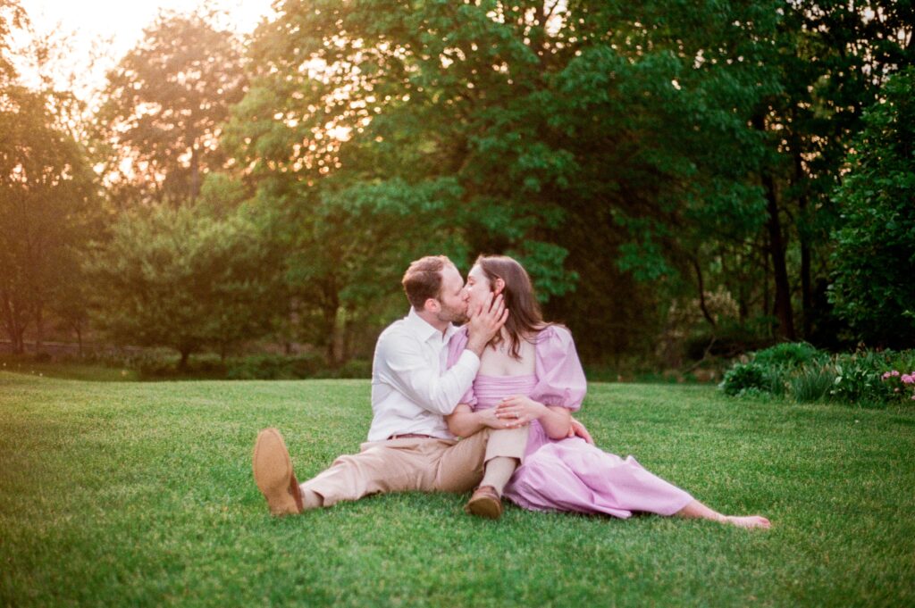 A couple sits in the grass and shares a kiss on film for their Northern Virginia engagement photographer