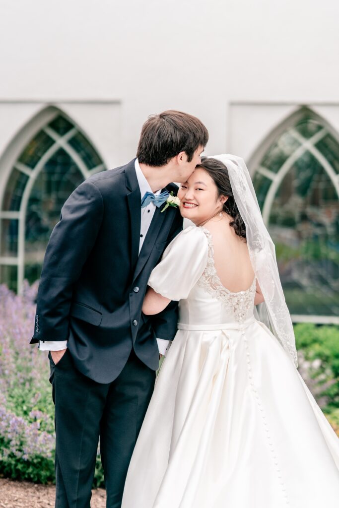 A bride smiles over her shoulder as her groom kisses her on the forehead by by Northern Virginia wedding photographer
