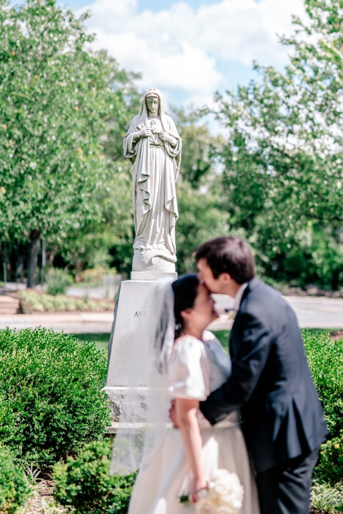 A groom kisses his bride on the forehead in front of a statue of Mary