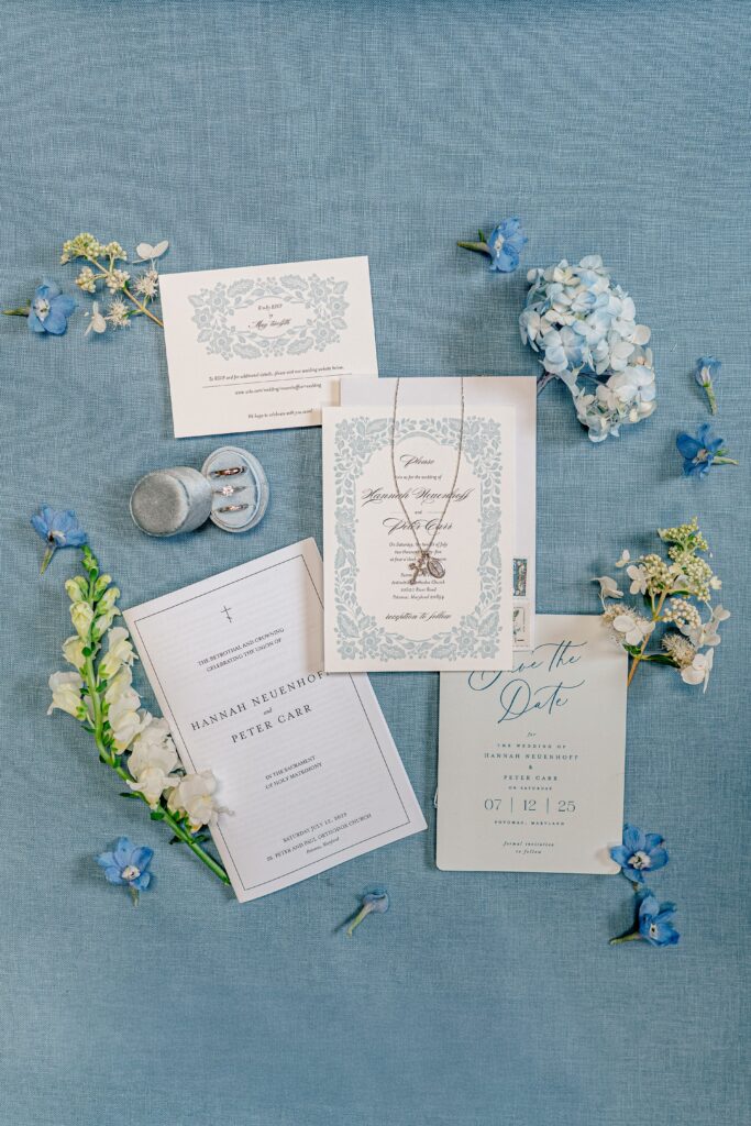 A wedding flat lay featuring a blue background, blue floral invitation suite, classic silver wedding rings, and blue hydrangea flowers