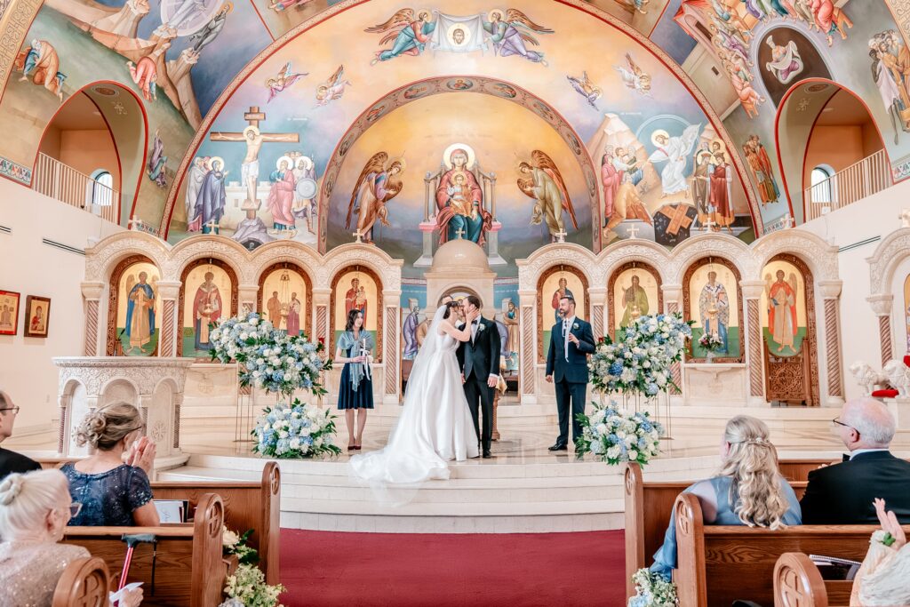A bride and groom sharing their first kiss during their wedding ceremony in a Greek Orthodox church