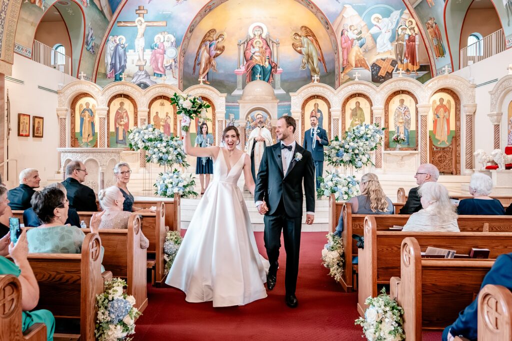 A bride and groom cheering as they walk down the aisle at the end of their Greek Orthodox wedding