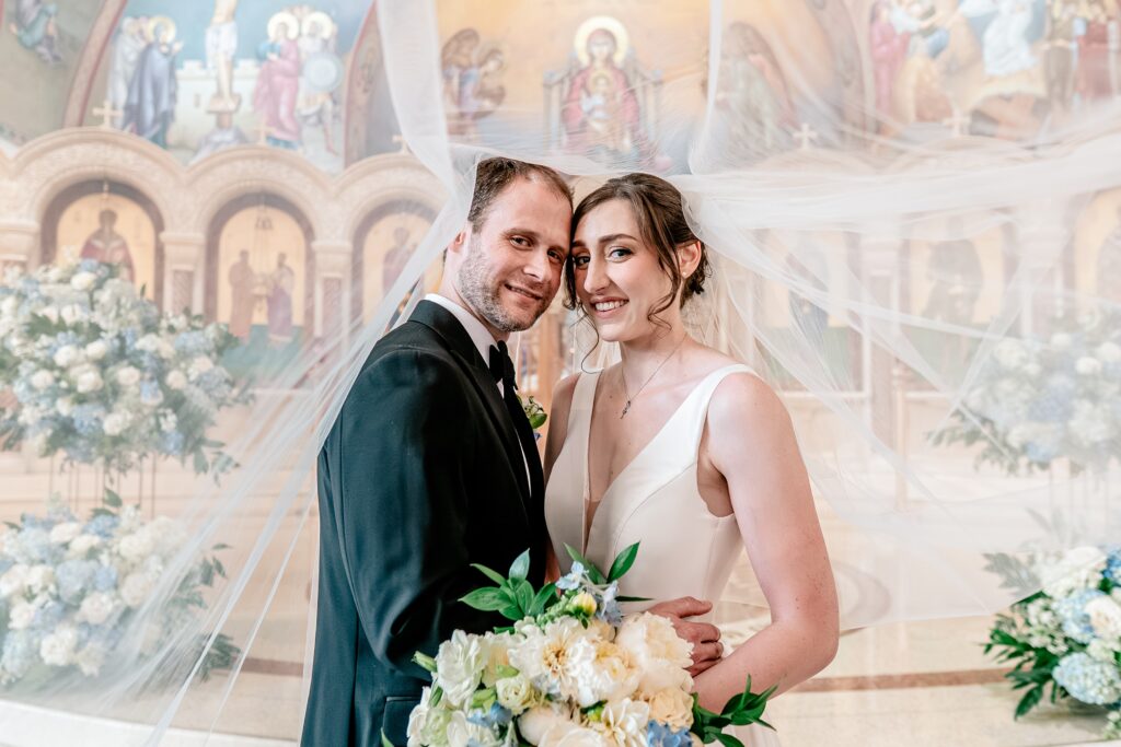 A bride and groom smiling at the camera with the veil around them during their Greek Orthodox wedding