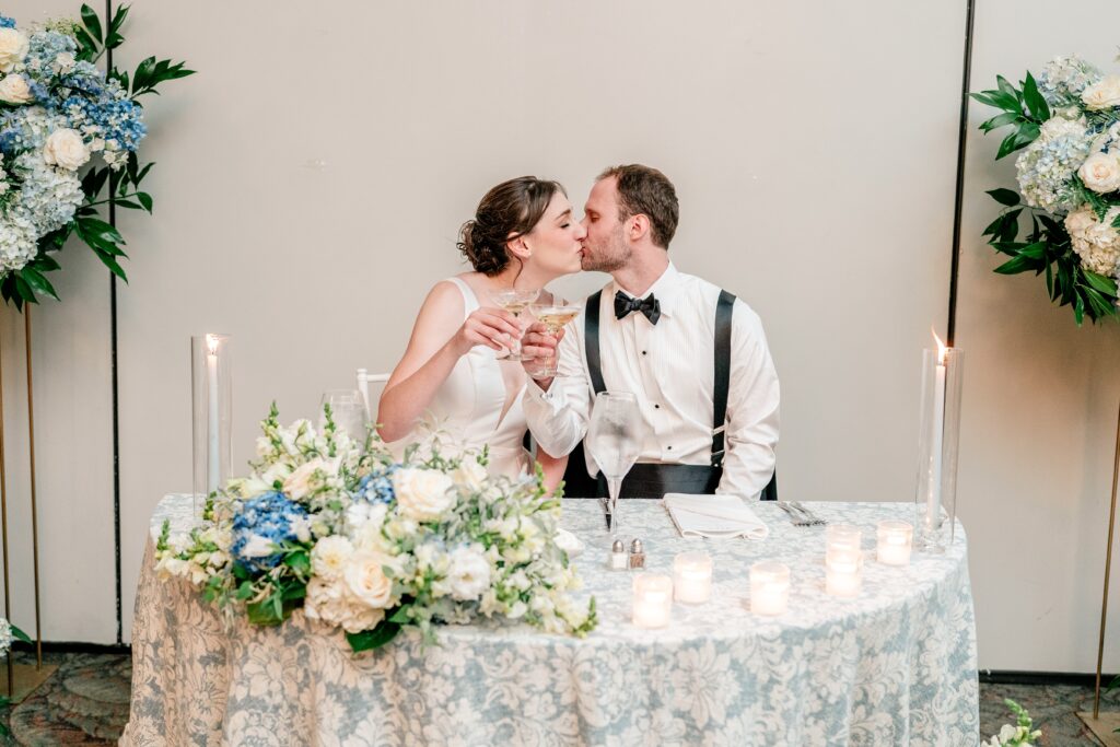 A bride and groom sharing a kiss while toasting their champagne coups at their sweetheart table