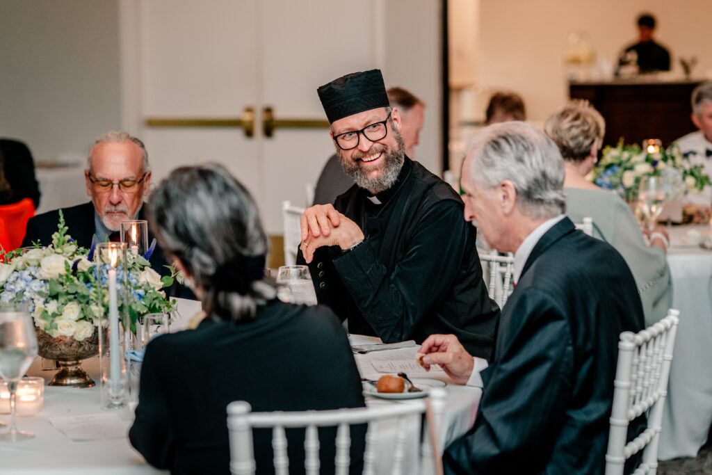 A priest laughing with wedding guests at a reception table