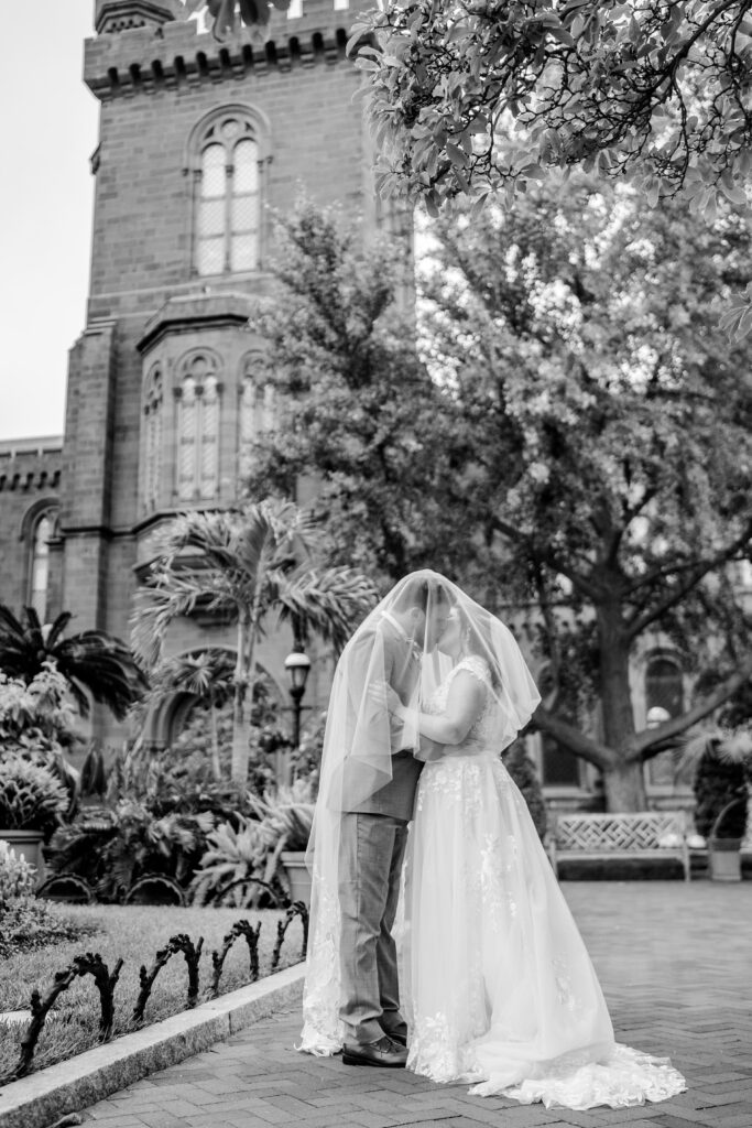 A black and white photo of a bride and groom sharing a kiss under the veil while standing in front of the Smithsonian Castle