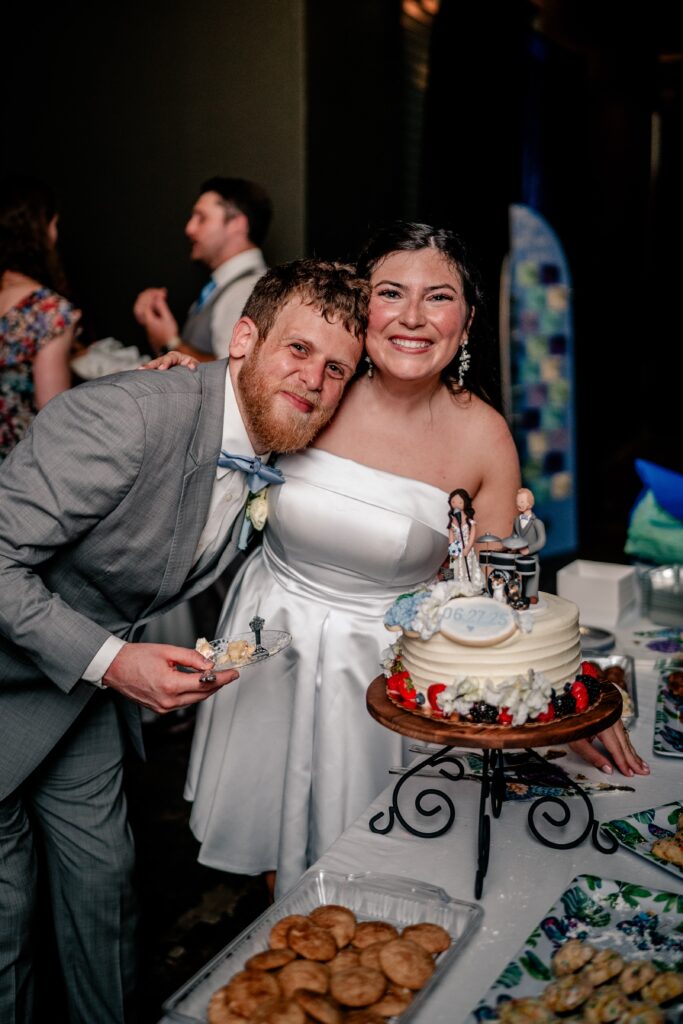 A bride and groom smiling at the camera while standing beside their wedding cake