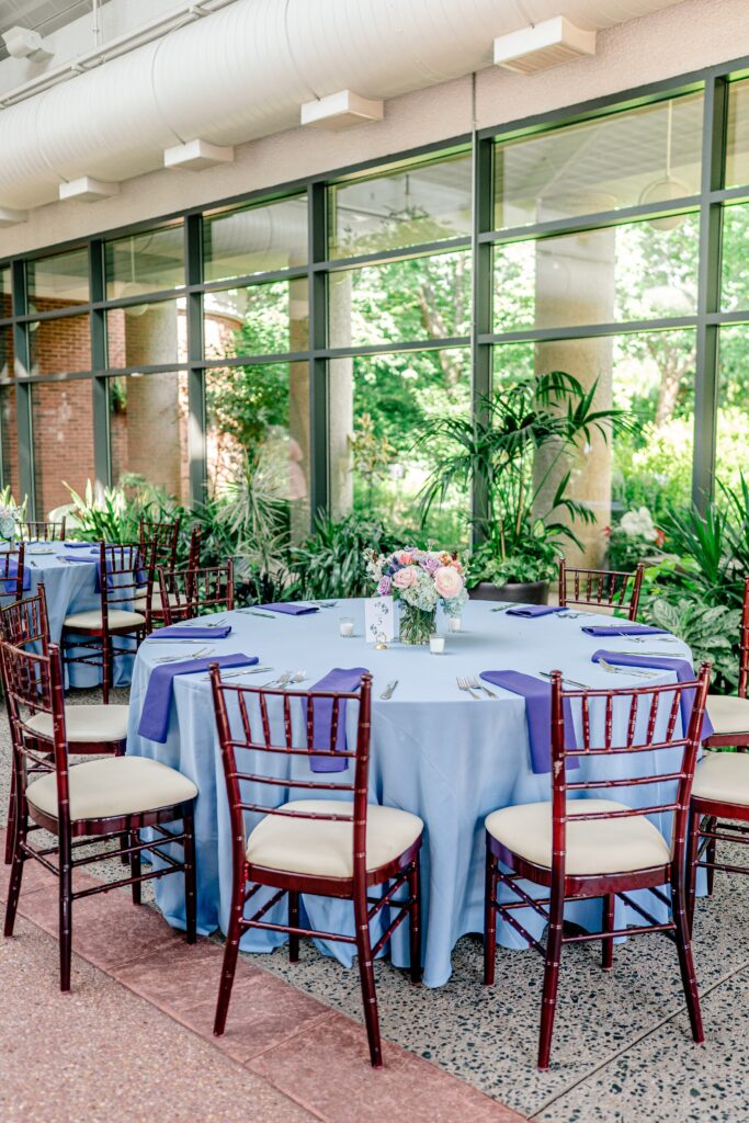 A table inside an atrium reception venue with a periwinkle tablecloth, wooden chiavari chairs, purple napkins, and a small colorful floral centerpiece