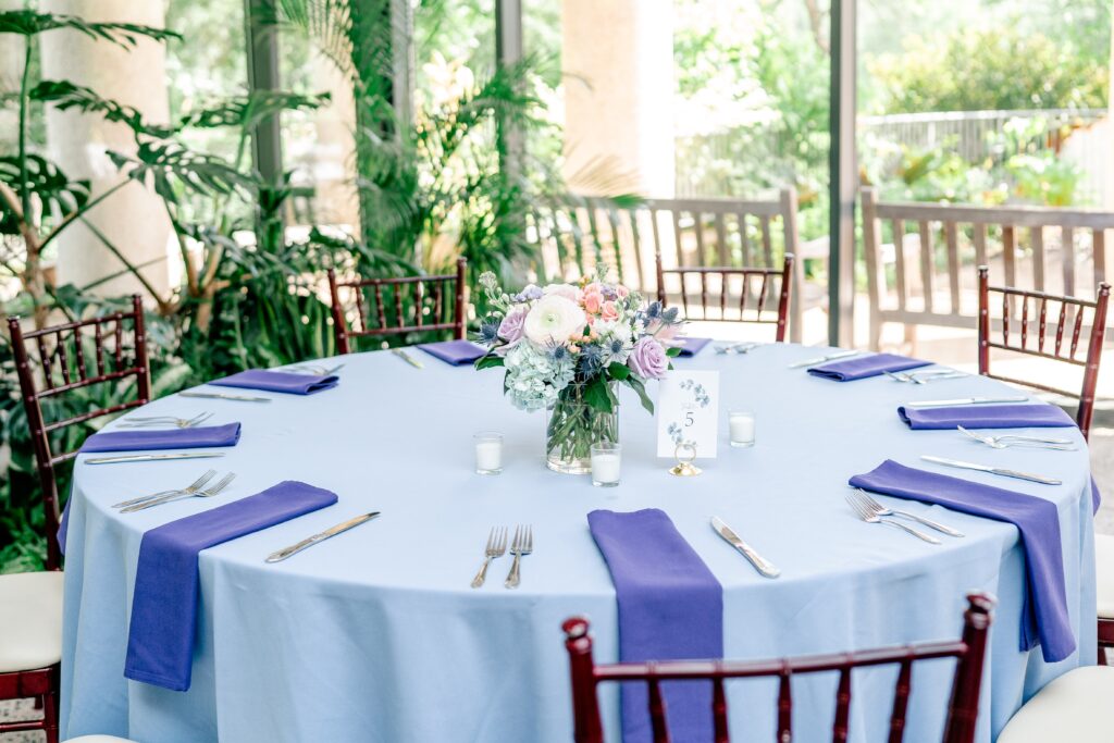 A beautiful flower arrangement on a reception table featuring purple napkins on a periwinkle tablecloth for a detail photo by a Northern Virginia wedding photographer