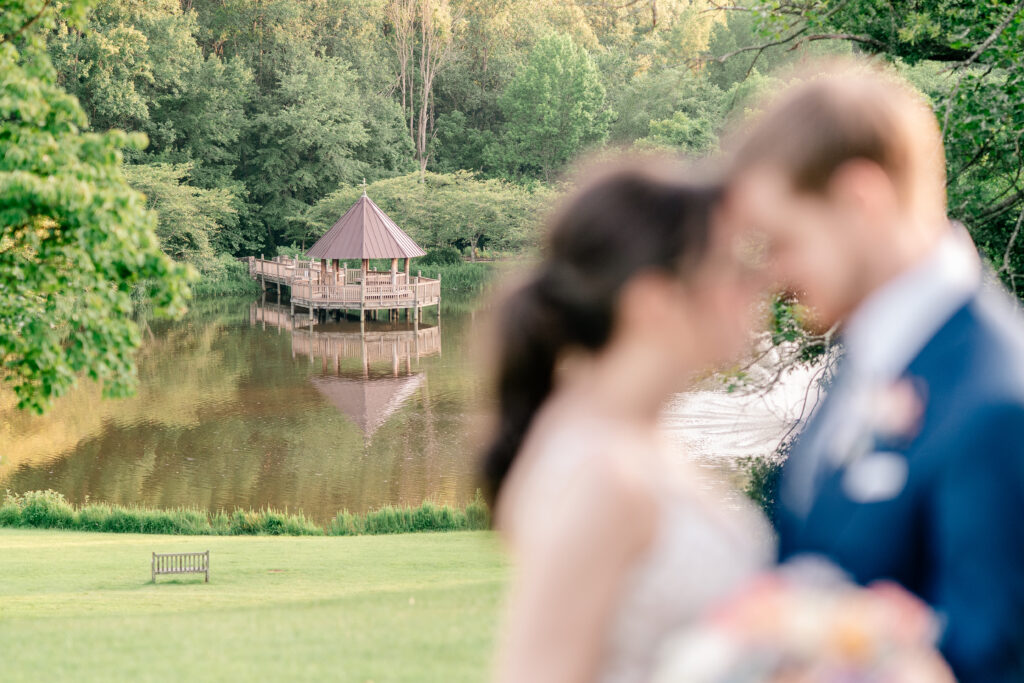 A gazebo on a lake off in the distance in focus while a bride and groom standing in the foreground out of focus