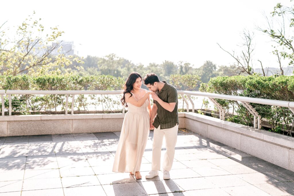 A man kissing the hand of his partner in bright sunlight standing on a terrace by the Potomac River