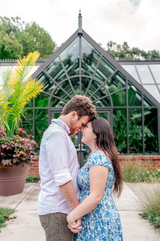 A couple stands forehead to forehead and holding hands in front of the greenhouse at Meadowlark Botanical Gardens