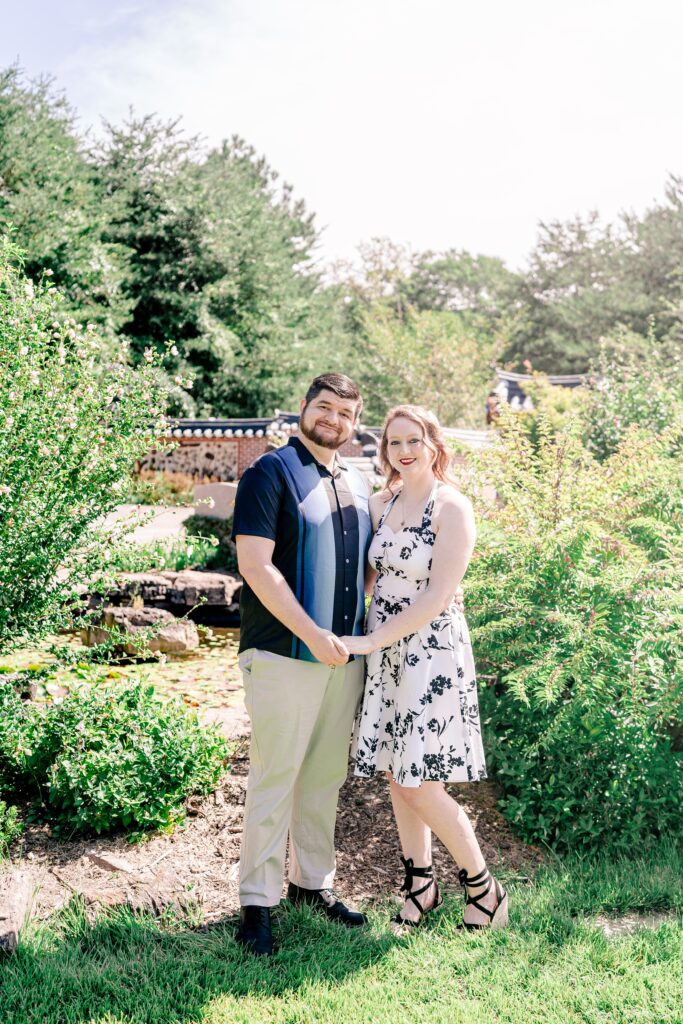 An engaged couple poses for a classic portrait on a sunny day in a garden with a small pond in the background