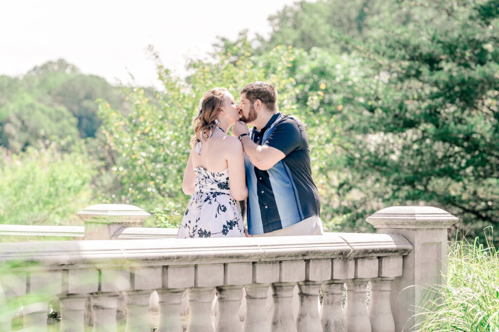 An engaged couple shares a kiss on a balcony surrounded by a stone railing