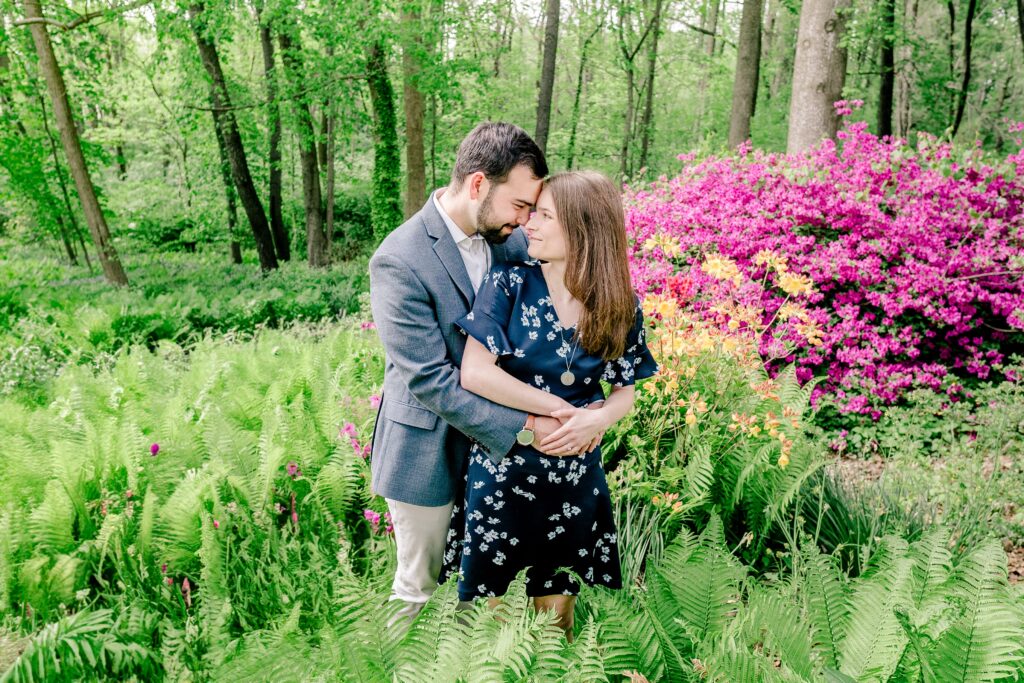 A man and woman nuzzle close together while standing in a garden with a vibrant purple bush behind them