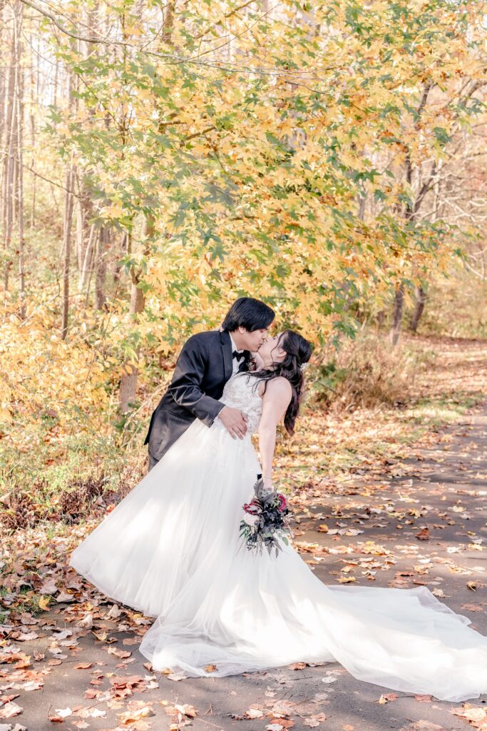 A bride and groom share a dip kiss with golden yellow leaves on the trees around them
